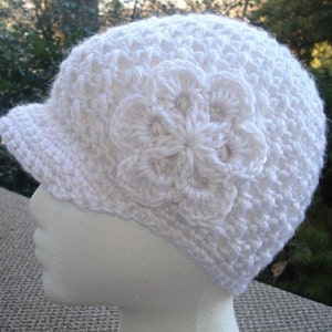 Puede incluir: Sombrero blanco de ganchillo con un diseño de flor en el lateral. El sombrero tiene una visera y está hecho con un punto texturizado.