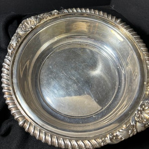 May include: A silver serving bowl with a decorative, rope-like rim and ornate floral designs on the sides. The bowl is polished and reflects light.
