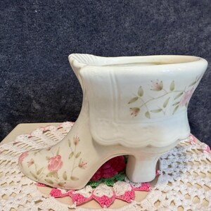 May include: A white ceramic shoe planter with a floral design featuring pink flowers and green leaves. The shoe has a high heel and a pointed toe.