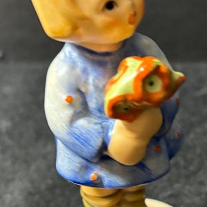 May include: A porcelain figurine of a young girl with blonde hair wearing a blue dress and holding a bouquet of red and green flowers. The figurine is standing on a cream-colored base.