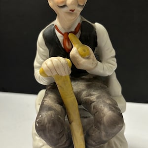 May include: A porcelain figurine of an elderly man with a mustache and a red tie, sitting on a white base with gold accents. He is holding a yellow cane and has a thoughtful expression on his face.