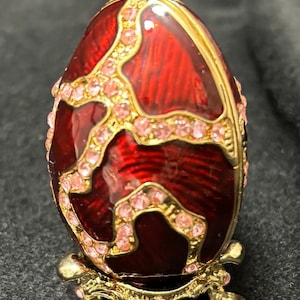 May include: A red and gold jeweled egg with pink rhinestones on a gold stand. The egg has a decorative design of swirls and lines.