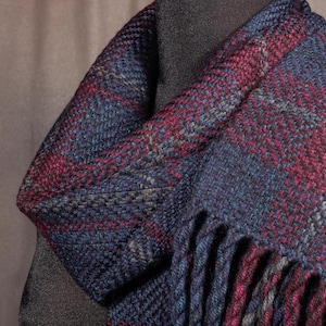 May include: A woven scarf featuring a textured herringbone pattern in deep shades of navy blue, burgundy, and charcoal gray. The fabric displays a dense, interlaced weave with visible yarn strands and a fringed edge with twisted tassels.