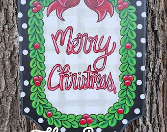 May include: A rectangular Christmas sign with a black border and white polka dots. It features a red bow, green garland, and the words "Merry Christmas" in red script. The sign is adorned with red berries and a beaded handle.