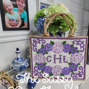 May include: A burlap tote bag featuring a purple floral design and the initials "CHL". The bag has a rectangular shape with a woven texture and brown handles. The text "The Sassy Paintbrush" is written in cursive.