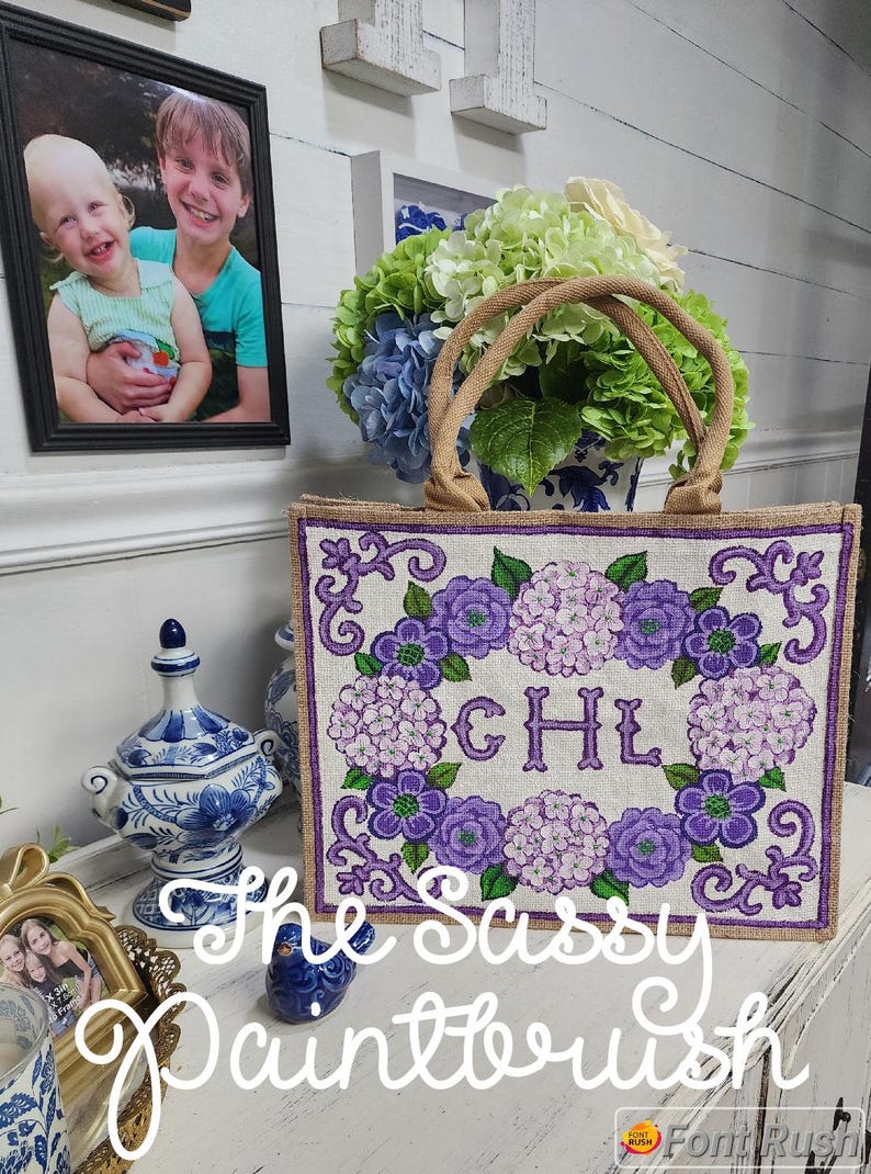 May include: A burlap tote bag featuring a purple floral design and the initials "CHL". The bag has a rectangular shape with a woven texture and brown handles. The text "The Sassy Paintbrush" is written in cursive.