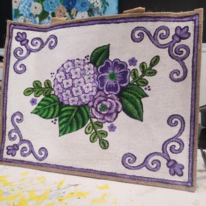May include: A rectangular tote bag with a floral design. The bag features a white background with a purple border and a painted bouquet of purple hydrangeas, roses, and green leaves. The bag has a burlap trim and handles.
