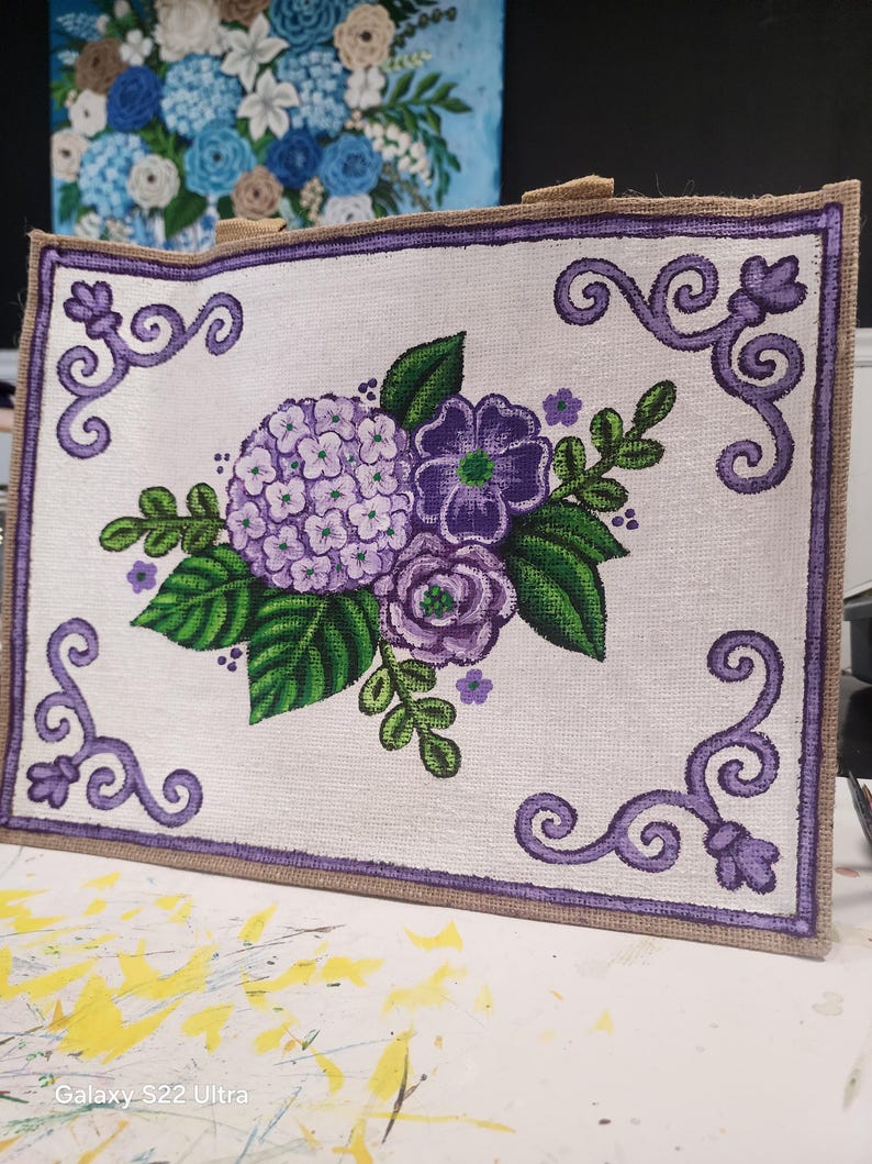 May include: A rectangular tote bag with a floral design. The bag features a white background with a purple border and a painted bouquet of purple hydrangeas, roses, and green leaves. The bag has a burlap trim and handles.