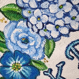 May include: Close-up of a floral painting featuring blue hydrangeas and other flowers with green leaves and gold accents. The flowers are painted on a white textured background. The painting has a decorative border.