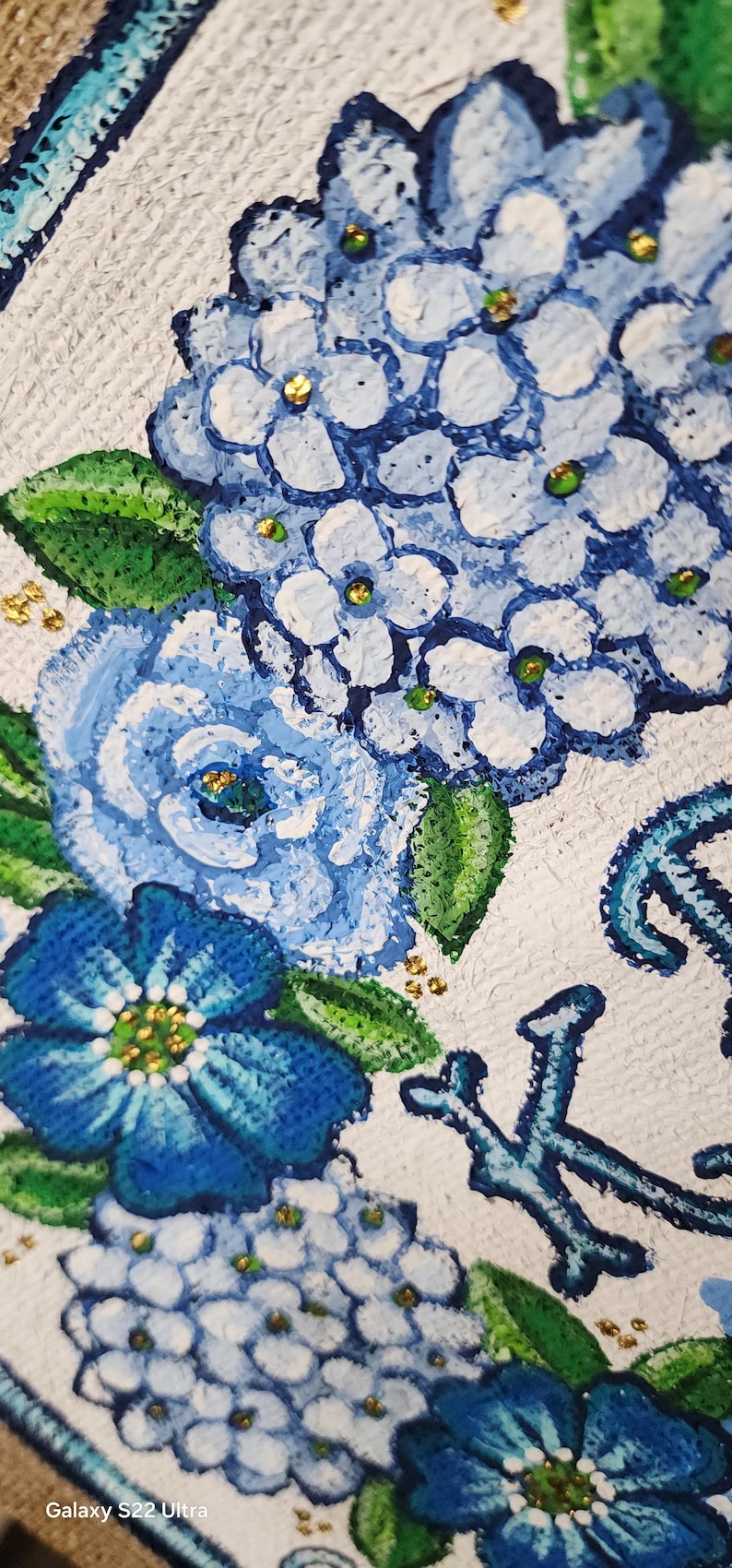 May include: Close-up of a floral painting featuring blue hydrangeas and other flowers with green leaves and gold accents. The flowers are painted on a white textured background. The painting has a decorative border.