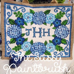 May include: A rectangular tote bag with a burlap exterior and a floral design. The central panel features blue hydrangeas and roses surrounding the monogram "JHH". The text "The Sassy Paintbrush" is written across the bottom.