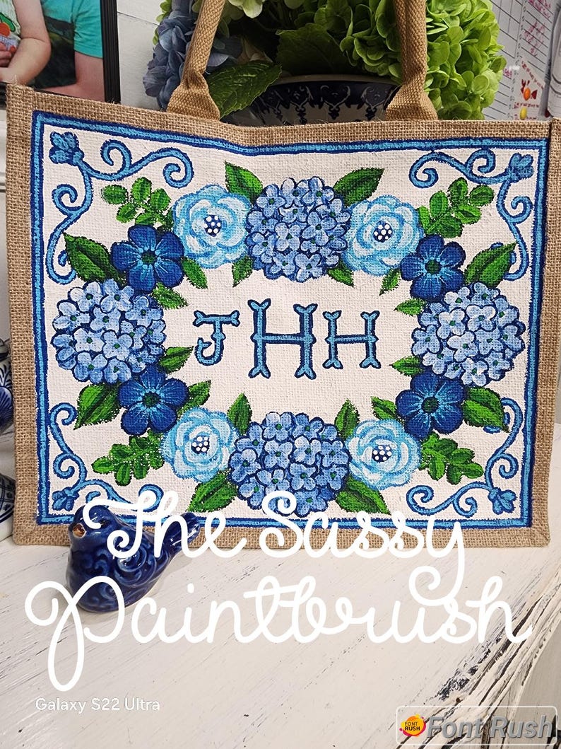 May include: A rectangular tote bag with a burlap exterior and a floral design. The central panel features blue hydrangeas and roses surrounding the monogram "JHH". The text "The Sassy Paintbrush" is written across the bottom.