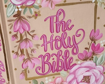 Pode incluir: Uma capa de livro bege com o título The Holy Bible em letra cursiva rosa escuro, emoldurada por flores pintadas à mão em rosa e branco com folhas verdes e detalhes dourados. A capa apresenta uma borda retangular dourada e motivos florais.
