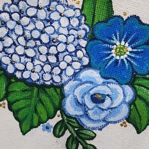 May include: Close-up of a floral painting featuring blue hydrangeas, a blue rose, and a blue flower with a yellow centre. The flowers are set against a white background with green leaves and gold accents. The painting has a rustic, handmade feel.
