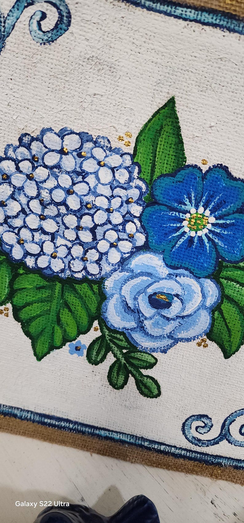May include: Close-up of a floral painting featuring blue hydrangeas, a blue rose, and a blue flower with a yellow centre. The flowers are set against a white background with green leaves and gold accents. The painting has a rustic, handmade feel.