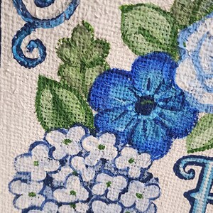May include: Close-up of a painting featuring blue flowers and green leaves on a white textured background. The flowers include hydrangeas and other blooms, with decorative blue swirls. The artwork is detailed and vibrant.
