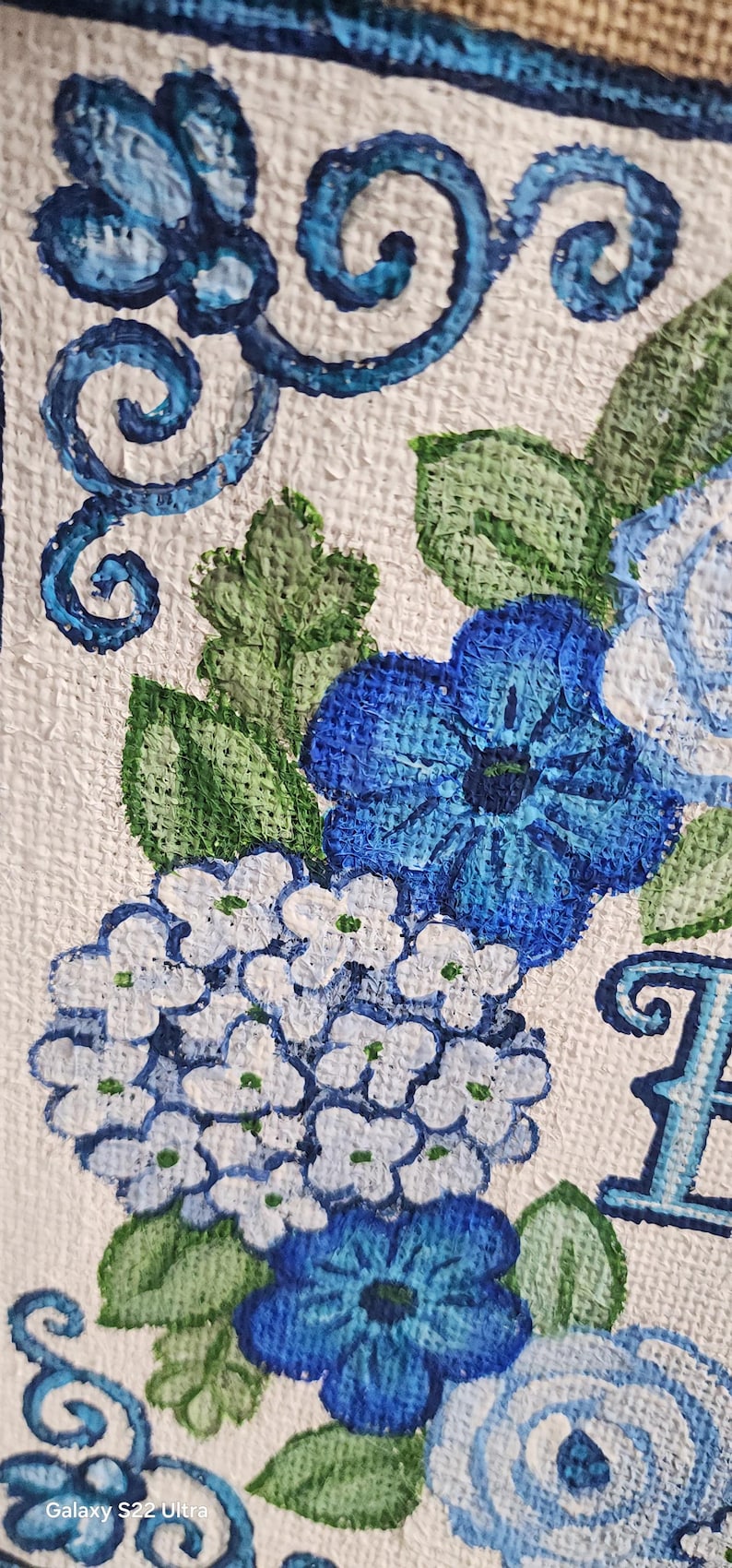 May include: Close-up of a painting featuring blue flowers and green leaves on a white textured background. The flowers include hydrangeas and other blooms, with decorative blue swirls. The artwork is detailed and vibrant.