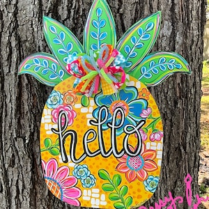 Può includere: Un appendiporta in legno a forma di ananas giallo con un motivo floreale e la parola "hello" in corsivo nero. L'ananas ha foglie verdi e un fiocco colorato.