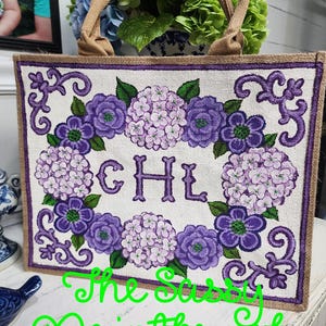 May include: A rectangular tote bag with a floral design featuring purple and lavender hydrangeas and the initials "CHL". The bag has a burlap-like texture, a brown border, and brown handles. The text "The Sassy Paintbrush" is written in green.