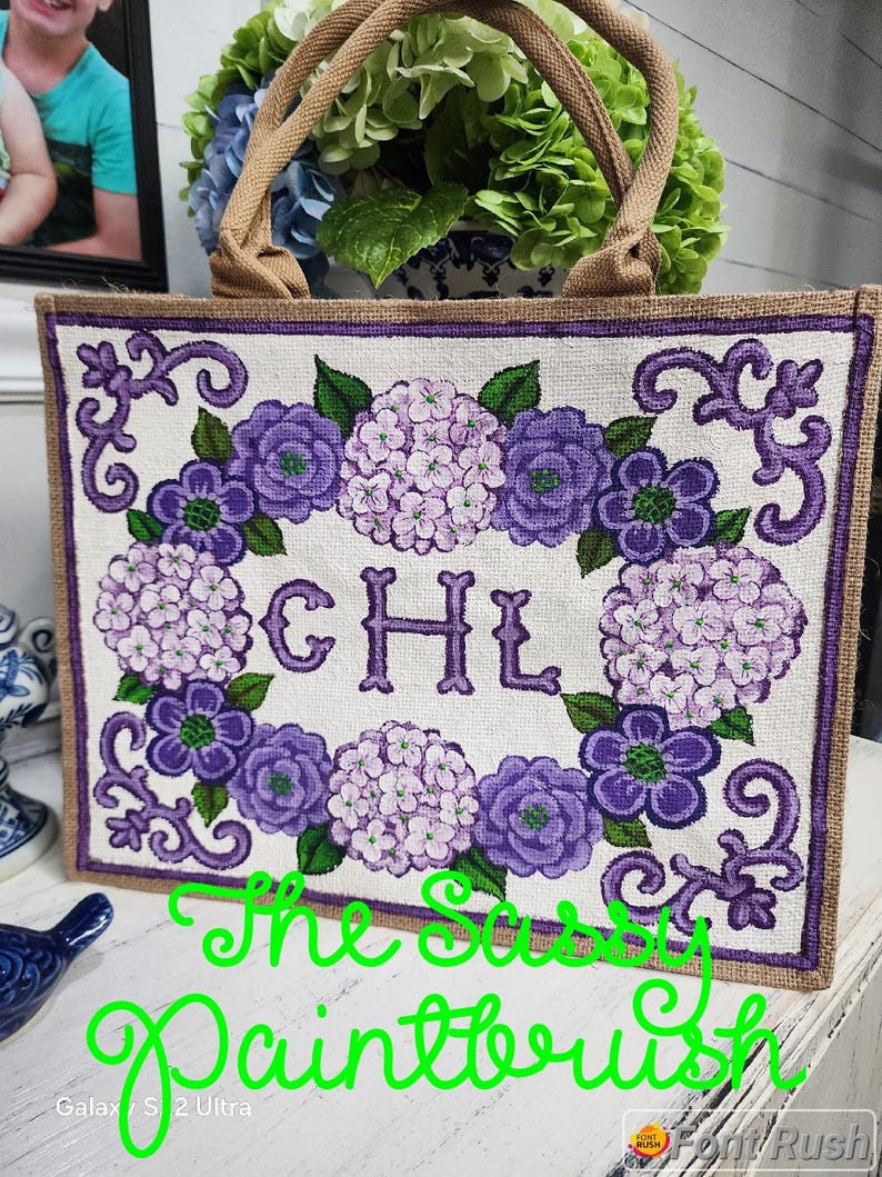 May include: A rectangular tote bag with a floral design featuring purple and lavender hydrangeas and the initials "CHL". The bag has a burlap-like texture, a brown border, and brown handles. The text "The Sassy Paintbrush" is written in green.