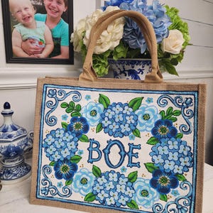 May include: A rectangular tote bag with a floral design featuring blue hydrangeas and the letters "BOE". The bag has a natural burlap exterior and a brown handle. The floral design is framed with a blue border.