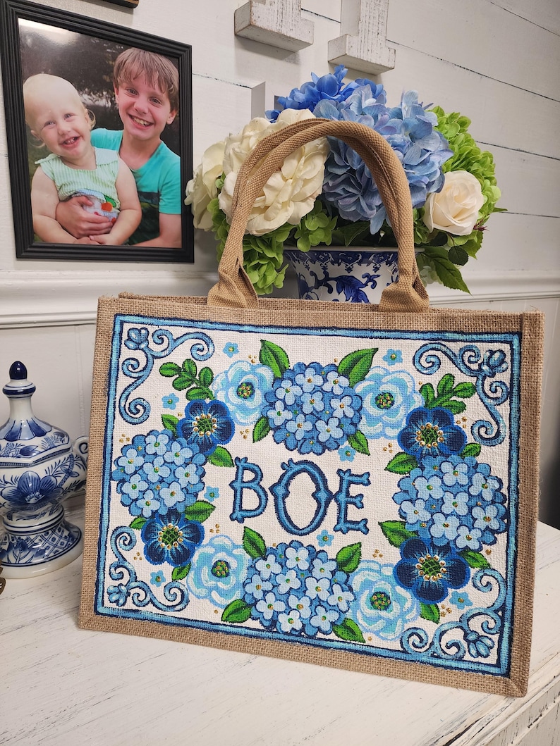 May include: A rectangular tote bag with a floral design featuring blue hydrangeas and the letters "BOE". The bag has a natural burlap exterior and a brown handle. The floral design is framed with a blue border.
