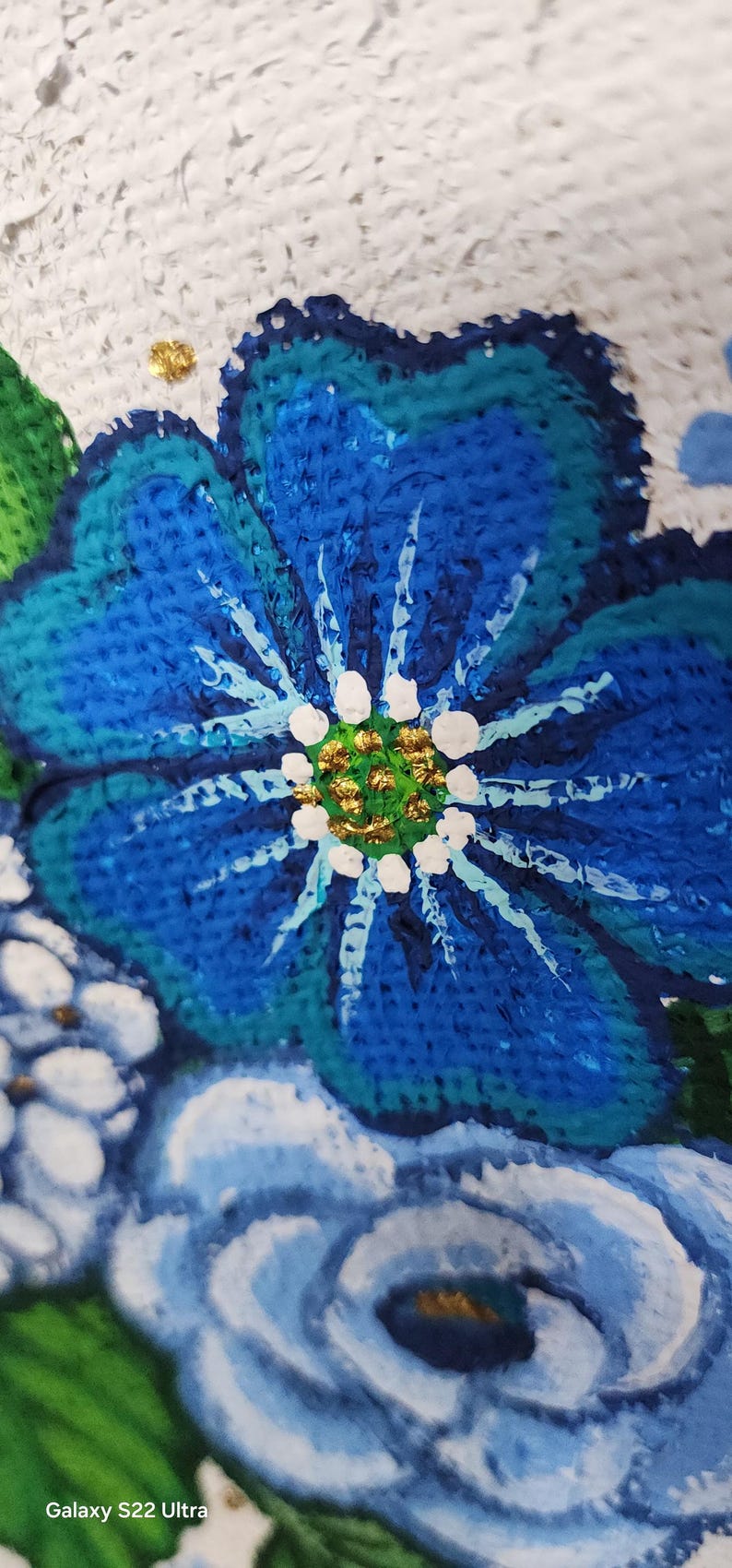 May include: Close-up of a vibrant, textured painting of a blue flower with white accents and a gold centre. The artwork features a textured white background and green leaves, showcasing a floral theme.