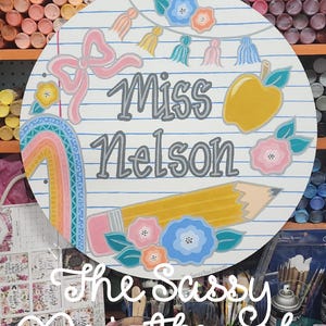 Könnte beinhalten: Runder Holzschild mit Schulthema. Es zeigt die Worte "Miss Nelson" in Grau, zusammen mit einem gelben Bleistift, Apfel und bunten Blumen. Eine rosa Schleife und ein Regenbogen sind ebenfalls enthalten. Das Schild hat einen Perlen-Griff.