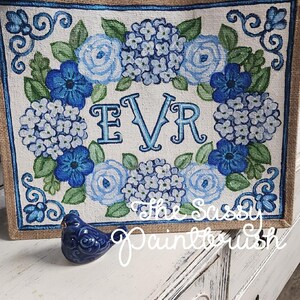 May include: A square burlap tote bag featuring a floral design in shades of blue and green. The design includes hydrangeas, roses, and the letters "EVR". The bag has a brown border and handles. A small blue bird figurine sits in front.