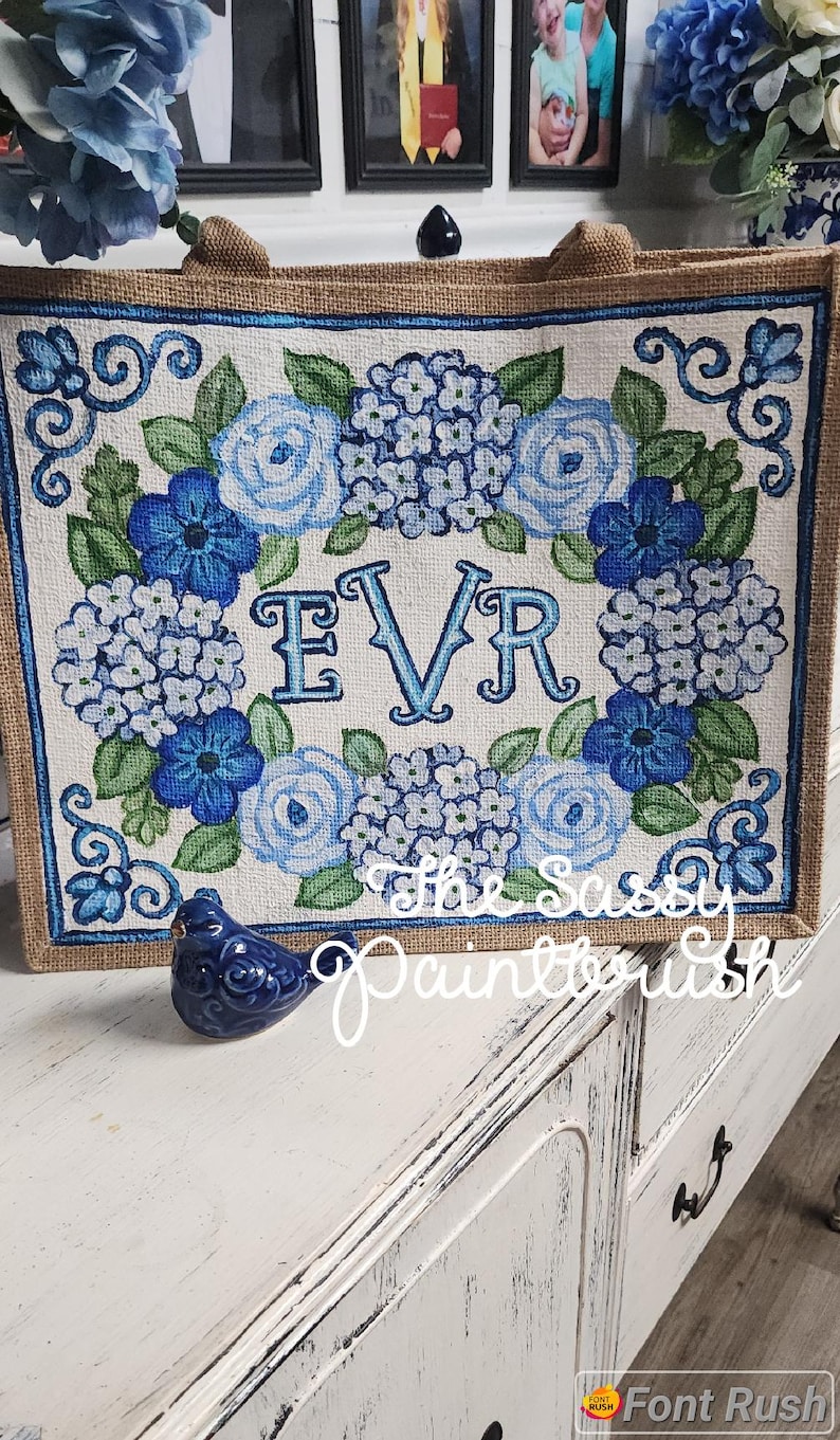 May include: A square burlap tote bag featuring a floral design in shades of blue and green. The design includes hydrangeas, roses, and the letters "EVR". The bag has a brown border and handles. A small blue bird figurine sits in front.