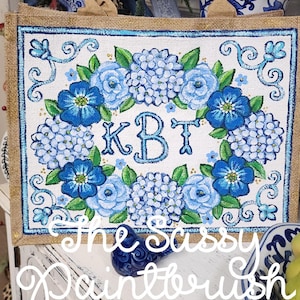 May include: A burlap tote bag with a floral design. The front of the bag features a white background with a blue floral wreath and the initials "KBT". The words "The Sassy Paintbrush" are written across the bottom.