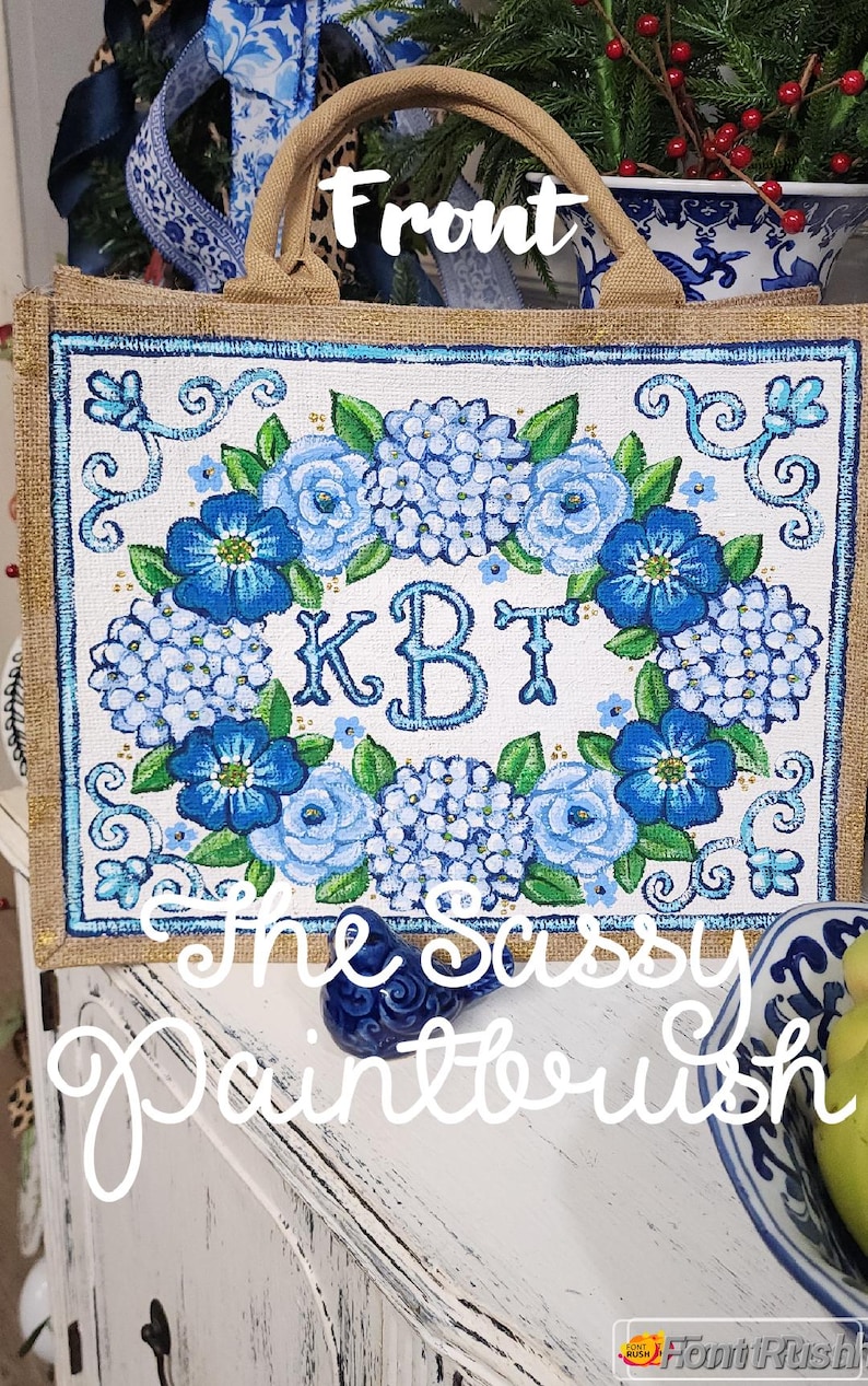 May include: A burlap tote bag with a floral design. The front of the bag features a white background with a blue floral wreath and the initials "KBT". The words "The Sassy Paintbrush" are written across the bottom.