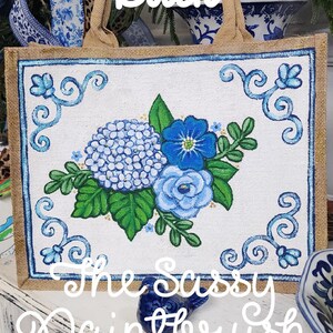 May include: A jute tote bag with a hand-painted floral design. The artwork features blue hydrangeas, roses, and other flowers, framed by a blue border. The text "Back" and "The Sassy Paintbrush" are visible.