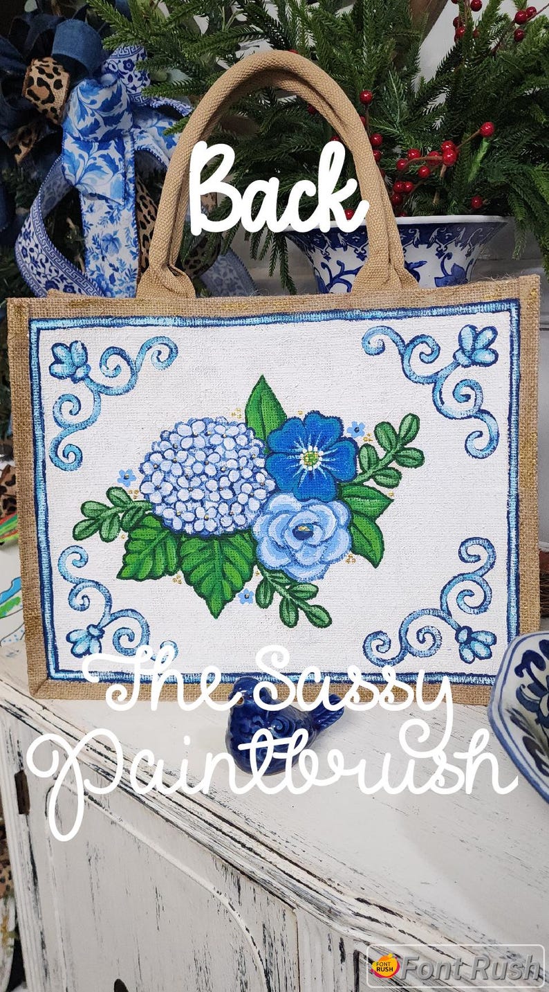 May include: A jute tote bag with a hand-painted floral design. The artwork features blue hydrangeas, roses, and other flowers, framed by a blue border. The text "Back" and "The Sassy Paintbrush" are visible.