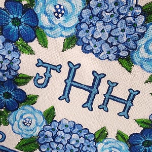 May include: A white canvas with a floral design featuring blue hydrangeas and other blue flowers with green leaves. The letters "THH" are painted in blue. The design is bordered with a blue stripe and burlap.