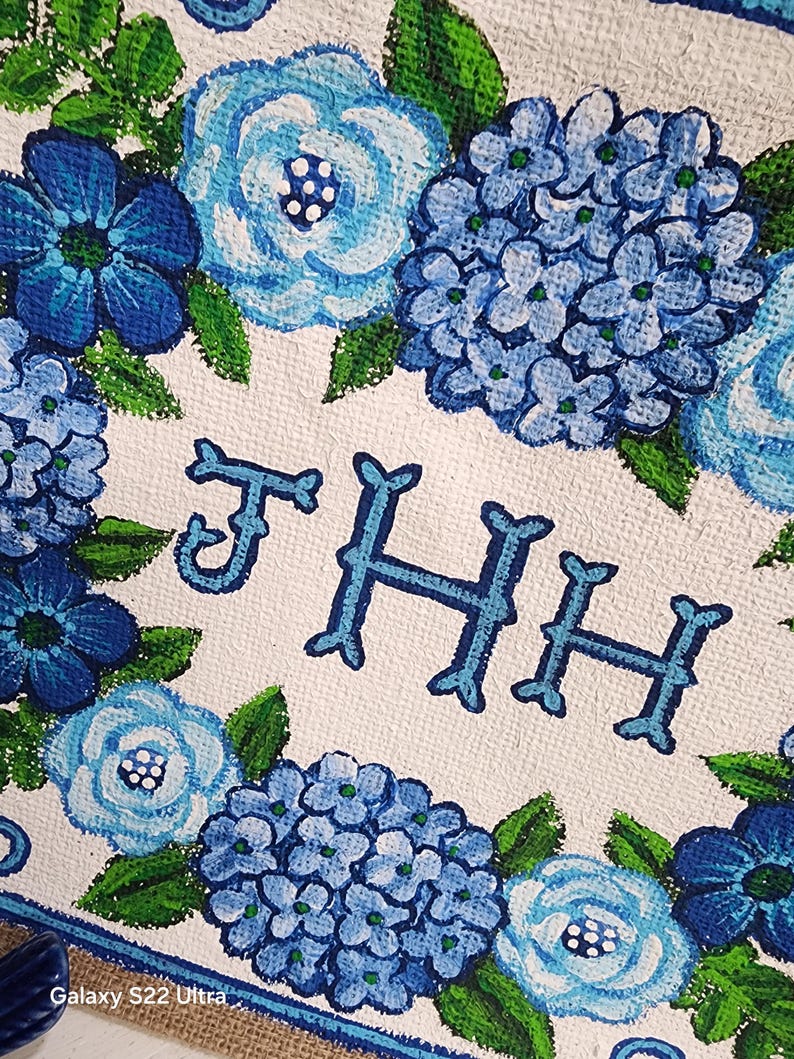 May include: A white canvas with a floral design featuring blue hydrangeas and other blue flowers with green leaves. The letters "THH" are painted in blue. The design is bordered with a blue stripe and burlap.