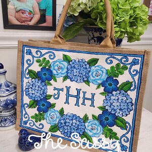 May include: A jute tote bag with a floral design of blue hydrangeas and roses. The bag has a beige handle and a blue border. The text "JHH" is in the centre of the floral design. The words "The Sassy Paintbrush" are written at the bottom.