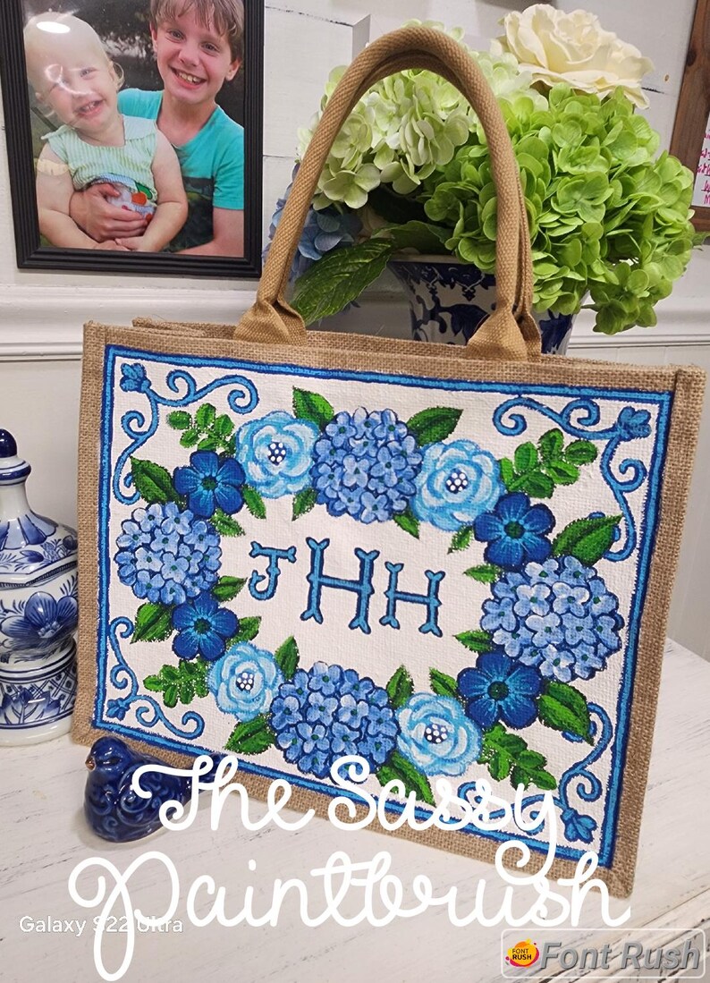 May include: A jute tote bag with a floral design of blue hydrangeas and roses. The bag has a beige handle and a blue border. The text "JHH" is in the centre of the floral design. The words "The Sassy Paintbrush" are written at the bottom.