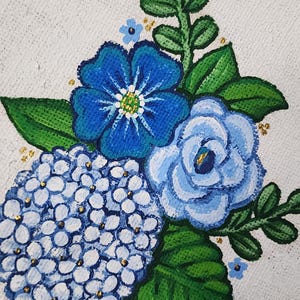May include: Hand-painted floral artwork featuring blue hydrangeas, roses, and other flowers with green leaves. The painting has a white background and a blue decorative border. Gold accents are scattered throughout the floral arrangement.