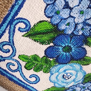 May include: A close-up of a hand-painted floral design featuring blue hydrangeas and other flowers with green leaves. The artwork is set against a white background with a blue border and a burlap edge. The painting is on a textured surface.
