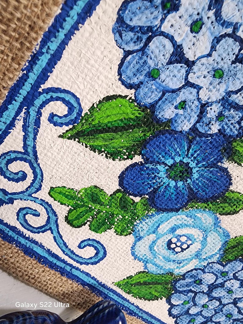 May include: A close-up of a hand-painted floral design featuring blue hydrangeas and other flowers with green leaves. The artwork is set against a white background with a blue border and a burlap edge. The painting is on a textured surface.