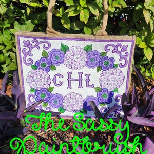 May include: A white tote bag with a floral design in shades of purple and green, featuring the initials "CHL". The bag has a burlap texture and sturdy handles. The text "The Sassy Paintbrush" is written in green cursive.