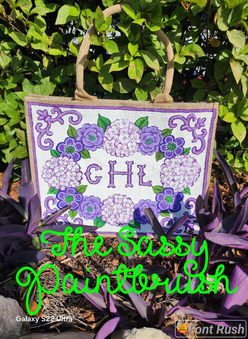 May include: A white tote bag with a floral design in shades of purple and green, featuring the initials "CHL". The bag has a burlap texture and sturdy handles. The text "The Sassy Paintbrush" is written in green cursive.