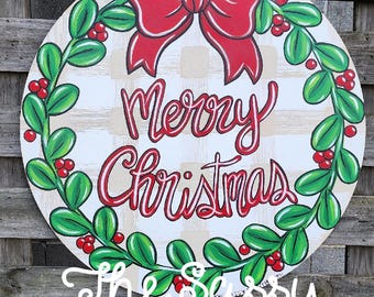 May include: Round wooden Christmas sign with a red bow, green leaves, and red berries. The sign says "Merry Christmas" in red script. The sign also says "The Sassy Paintbrush" in white script.