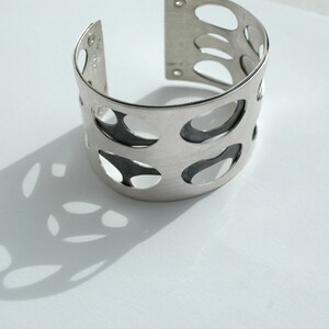 Puede incluir: Un brazalete plateado con un dise&ntilde;o calado. La pulsera presenta una serie de cortes ovalados, creando un patr&oacute;n de espacio negativo. El brazalete est&aacute; sobre una superficie blanca, proyectando una sombra.
