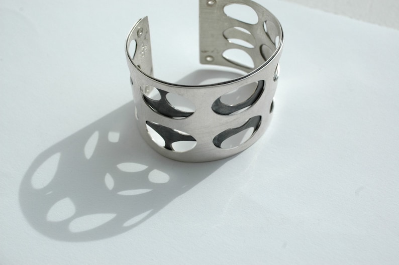 Puede incluir: Un brazalete plateado con un dise&ntilde;o calado. La pulsera presenta una serie de cortes ovalados, creando un patr&oacute;n de espacio negativo. El brazalete est&aacute; sobre una superficie blanca, proyectando una sombra.