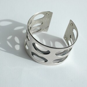 Puede incluir: Un brazalete plateado con un dise&ntilde;o calado. El brazalete tiene forma curva y presenta m&uacute;ltiples cortes ovalados. La imagen est&aacute; tomada sobre una superficie blanca, con el brazalete proyectando una sombra.