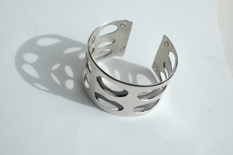 Puede incluir: Un brazalete plateado con un dise&ntilde;o calado. El brazalete tiene forma curva y presenta m&uacute;ltiples cortes ovalados. La imagen est&aacute; tomada sobre una superficie blanca, con el brazalete proyectando una sombra.