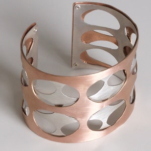 Puede incluir: Un brazalete ancho con un dise&ntilde;o de dos tonos. El brazalete presenta un exterior de color cobre con una serie de recortes ovalados, que revelan un interior de color plateado. El dise&ntilde;o abierto a&ntilde;ade un toque moderno a esta pieza de joyer&iacute;a.