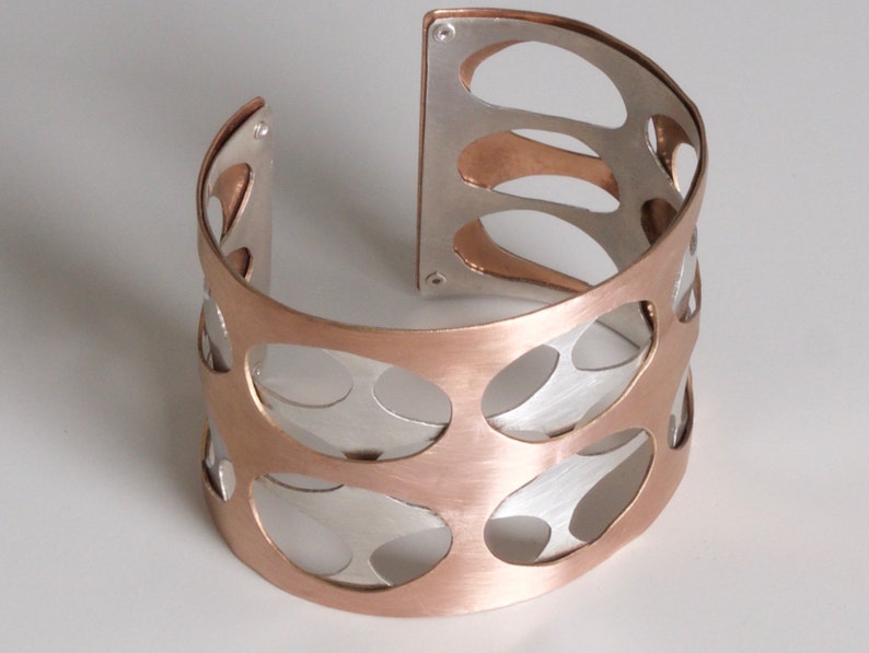 Puede incluir: Un brazalete ancho con un dise&ntilde;o de dos tonos. El brazalete presenta un exterior de color cobre con una serie de recortes ovalados, que revelan un interior de color plateado. El dise&ntilde;o abierto a&ntilde;ade un toque moderno a esta pieza de joyer&iacute;a.